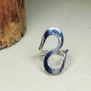 Peut inclure: Une bague en argent poli présentant un design ouvert et fluide en forme de S qui se courbe verticalement. La surface métallique est lisse et réfléchissante, créant une boucle continue et allongée qui se tient droite.