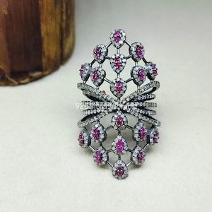 Puede incluir: Un anillo ancho y alargado de metal oscuro, con un diseño de celosía adornado con numerosas piedras blancas pequeñas y redondas y piedras rosas en forma de lágrima. El centro del anillo muestra el texto 925SilverVictoria.