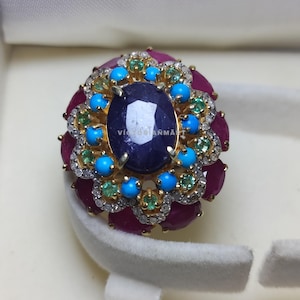 May include: A gold ring with a large, oval, dark blue gemstone at its centre. The ring is encircled by smaller, round, ruby-coloured stones, turquoise stones, and emerald stones, with small clear stones accenting the design.