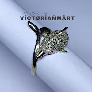 May include: Silver-toned ring featuring a dolphin design. The dolphin's body is encrusted with small, sparkling stones, and it has a red gemstone eye. The ring is displayed on a white surface, with the text "JAIPUR VICTORIANMART" visible.