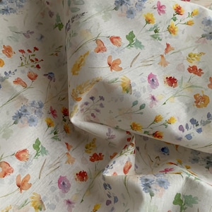 Puede incluir: Tela blanca con un estampado floral en acuarela. El diseño presenta una variedad de flores en tonos naranja, amarillo, morado y azul, con hojas y tallos verdes. La tela tiene un aspecto suave y fluido.