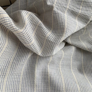May include: Close-up of a folded fabric with a striped pattern. The fabric has a natural, off-white base colour with thin, vertical stripes in a muted blue. The fabric is soft and appears to be linen or a similar material.