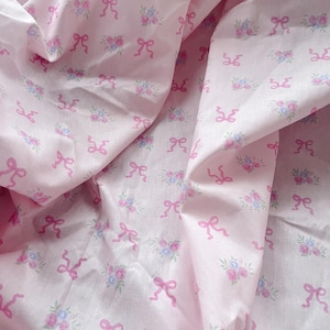 Op de afbeelding: Zachtroze stof met een herhaald patroon van roze strikken en kleine bloemboeketten in roze, blauw en groen. De stof is licht gekreukt. Het ontwerp heeft een delicate, vrouwelijke uitstraling.