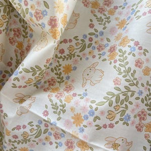 Può includere: Un tessuto bianco con un motivo ripetitivo di piccoli conigli dai colori chiari, intervallati da delicati motivi floreali in tonalità rosa tenue, giallo e azzurro, accompagnati da sottili steli e foglie verdi in tutto il design.
