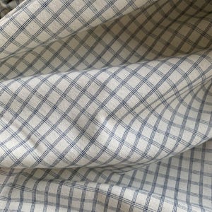 May include: Close-up of white fabric with a blue check pattern. The fabric is folded, creating shadows and depth. The check design is a classic pattern, suitable for various sewing projects or home decor.