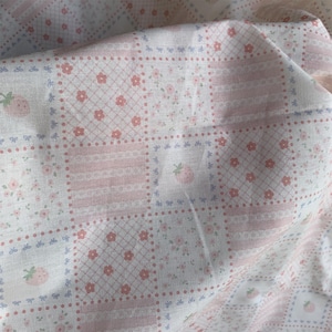 May include: A folded fabric with a patchwork design. The fabric features squares with floral, strawberry, and bow patterns in pink, white, and light blue. The fabric has a soft, textured appearance.