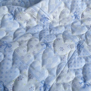 May include: A quilted fabric with a repeating heart pattern. The hearts are outlined with white stitching and filled with various blue and white patterns, including polka dots, floral prints, and checkered designs. Blue bows are placed throughout the design.