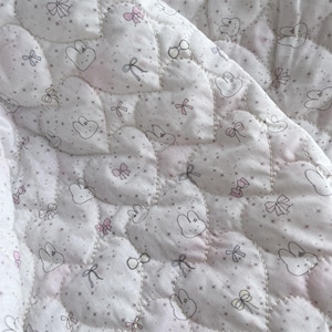 May include: Close-up of a quilted baby blanket with a repeating pattern of heart-shaped designs. The hearts are filled with bunny faces, bows, and small stars. The blanket is primarily white with pink and gray accents.