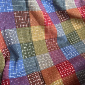 Puede incluir: Primer plano de una tela de patchwork con un patrón de cuadrícula. La tela presenta cuadrados de varios colores, incluyendo azul, rojo, morado, verde y marrón. Las costuras blancas delinean cada cuadrado, creando un efecto texturizado. La tela está ligeramente arrugada.