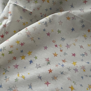 Può includere: Tessuto bianco con un motivo di stelle multicolore in tonalità giallo, rosa, blu e verde. Le stelle variano in dimensioni e sono stampate sul tessuto. Il tessuto ha una consistenza leggermente stropicciata.
