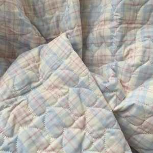 Peut inclure: Une couverture matelassée à carreaux roses et bleus pastel. La couverture présente un motif de matelassage en forme de cœur. Le tissu est doux et semble être du coton ou une matière similaire. La couverture est pliée, montrant le motif et la texture.