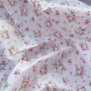 以下が含まれることがあります： 白い生地に繊細な花柄。ピンクと赤の花と緑の葉のクラスターが生地全体に散らばっています。生地は柔らかく、わずかにしわが寄った外観で、軽い質感を示唆しています。