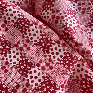 May include: Close-up of a patchwork fabric featuring a repeating pattern of red apples, white flowers, and red gingham checks on a white background. The fabric is folded, showing the texture and design variations.