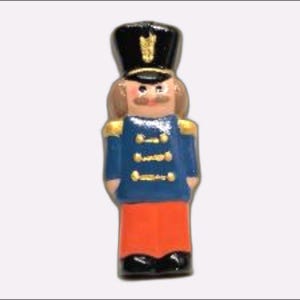 May include: A miniature ceramic nutcracker figurine. The figurine is dressed in a blue jacket with gold accents, orange pants, and a black hat. The nutcracker has a brown mustache and hair.