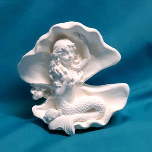 May include: White ceramic figurine of a mermaid sitting inside a clam shell. The mermaid has long hair and a tail.