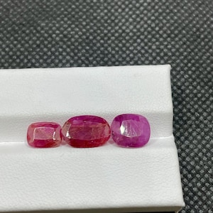 以下が含まれることがあります： さまざまなサイズの3つのファセットルビーが白い表面に展示されています。宝石は楕円形とクッションカットで、深い赤色を示し、内部構造と光の反射が見えます。ルビーは暗く、質感のある背景に配置されています。