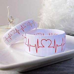 May include: Two white paper bracelets with a red heart beat design. The bracelets are on a white plate.