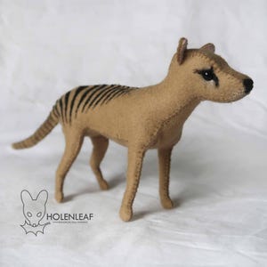 Può includere: Un giocattolo in feltro marrone che rappresenta un tilacino, noto anche come tigre della Tasmania, con strisce nere sulla schiena. Il giocattolo ha una lunga coda, piccole orecchie e un naso nero. Il logo "Holenleaf" è in basso a sinistra.