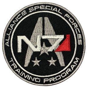 May include: A black and white embroidered patch with the text "Alliance Special Forces Training Program" around the edge. The center of the patch features a stylized "NZ" with three stars and a red triangle.