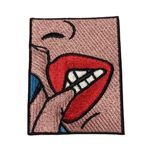 May include: Embroidered iron-on patch featuring a cartoon face with a wide open mouth, red lips, and white teeth. The face is pink and the background is a light pink.