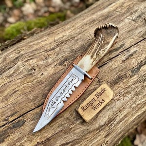 Könnte beinhalten: Handgefertigtes Jagdmesser mit gezahnter Klinge und Hirschhorngriff. Die Klinge hat geätzten Text und ist in einer Lederscheide untergebracht. Ein Holzschildchen trägt die Aufschrift "Ranger Blade Works".