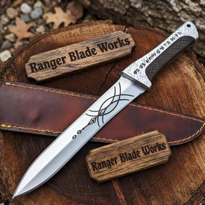 May include: A silver blade knife with a decorative handle and etched design, resting on a brown leather sheath. The knife has a dark handle and is accompanied by two wooden plaques that read "Ranger Blade Works". The knife is displayed on a wooden surface.