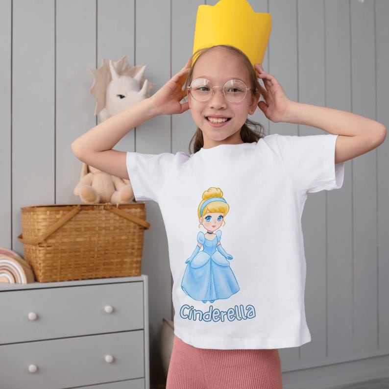 Peut inclure: Un t-shirt blanc avec une image de dessin anim&eacute; de Cendrillon dans une robe bleue. Le texte "Cendrillon" est imprim&eacute; sous l'image. Une jeune personne porte le t-shirt et une couronne jaune.
