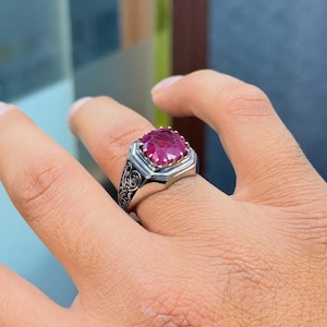 May include: A silver ring with a large, square-cut, deep red gemstone. The ring has a detailed, ornate design on the band and a geometric setting around the stone. The gemstone is the focal point, catching the light and showcasing its rich color.