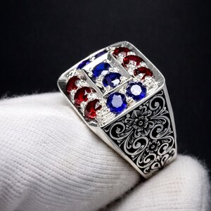 Puede incluir: Un anillo de plata con una cara rectangular adornada con piedras preciosas rojas y azules. La cara del anillo está dividida en secciones, cada una con múltiples piedras preciosas. La banda presenta intrincados grabados florales.