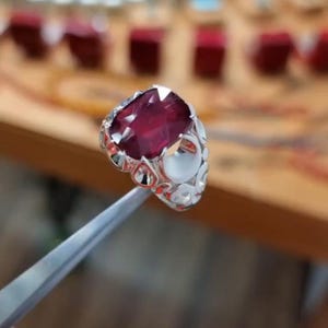 May include: A silver ring with a large, oval-cut, deep red gemstone. The ring's band features an ornate, openwork design with small, round accents. The ring is held by a pair of silver tweezers, with a blurred background.