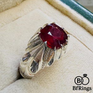 Könnte beinhalten: Ein silberner Ring mit einem großen, facettierten, tiefroten Edelstein. Der Ring hat ein verziertes Design mit detaillierten Metallarbeiten um den Stein herum. Der Ring ist vor einem neutralen Hintergrund platziert. Das Logo "BfRings" ist sichtbar.