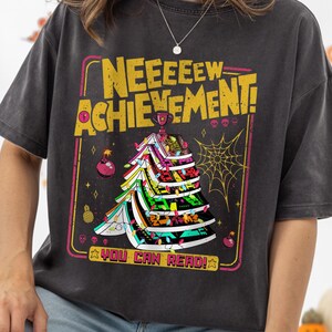 May include: Dark gray t-shirt with a colorful graphic that reads "NEEEEEEW ACHIEVEMENT!" and "YOU CAN READ!" in yellow and pink. The graphic features a decorated tree with a spider web and ornaments. The shirt is a relaxed fit.