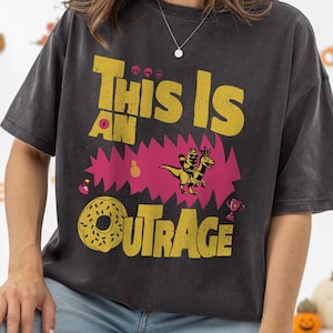 May include: Dark gray t-shirt with the text "This Is An Outrage" in yellow, featuring a pink jagged design and a cartoon dinosaur. The shirt has a vintage, distressed look, suitable for Halloween.
