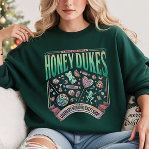 May include: A dark green sweatshirt featuring a colorful graphic of candies and the text "HONEYDUKES" and "LEGENDARY WIZARDING SWEET SHOP". The design includes lollipops, gummy bears, and other sweets.