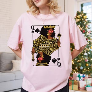 May include: A pale pink t-shirt with a Queen of Spades playing card design. The card shows a Black woman wearing a crown and sunglasses, with gold and black geometric patterns. The t-shirt is a relaxed fit.