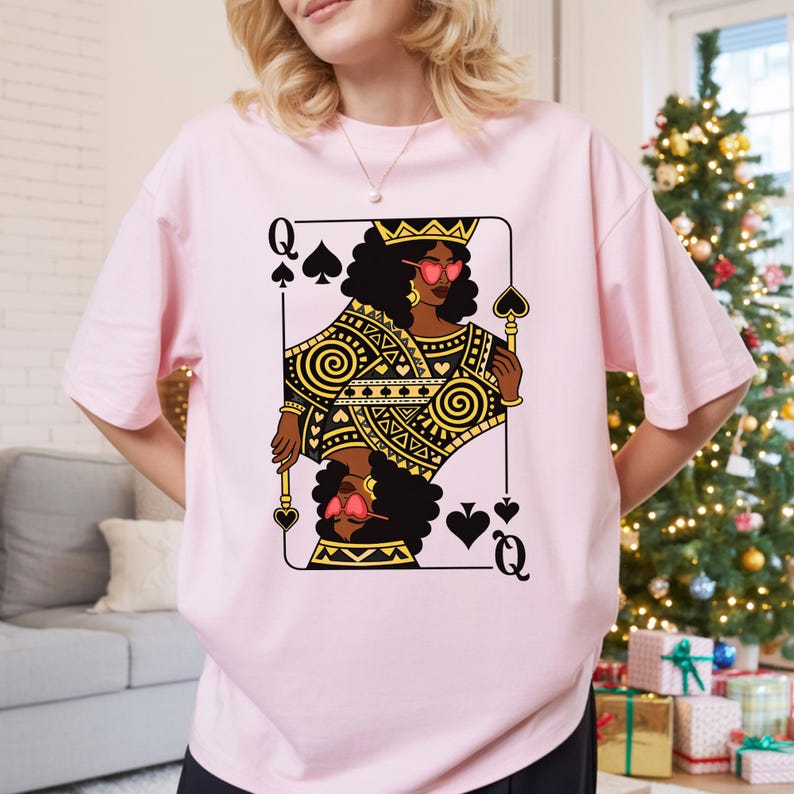 May include: A pale pink t-shirt with a Queen of Spades playing card design. The card shows a Black woman wearing a crown and sunglasses, with gold and black geometric patterns. The t-shirt is a relaxed fit.