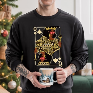 May include: Black sweatshirt with a gold and black playing card design featuring a queen wearing sunglasses. The person is holding a white mug with a winter scene.