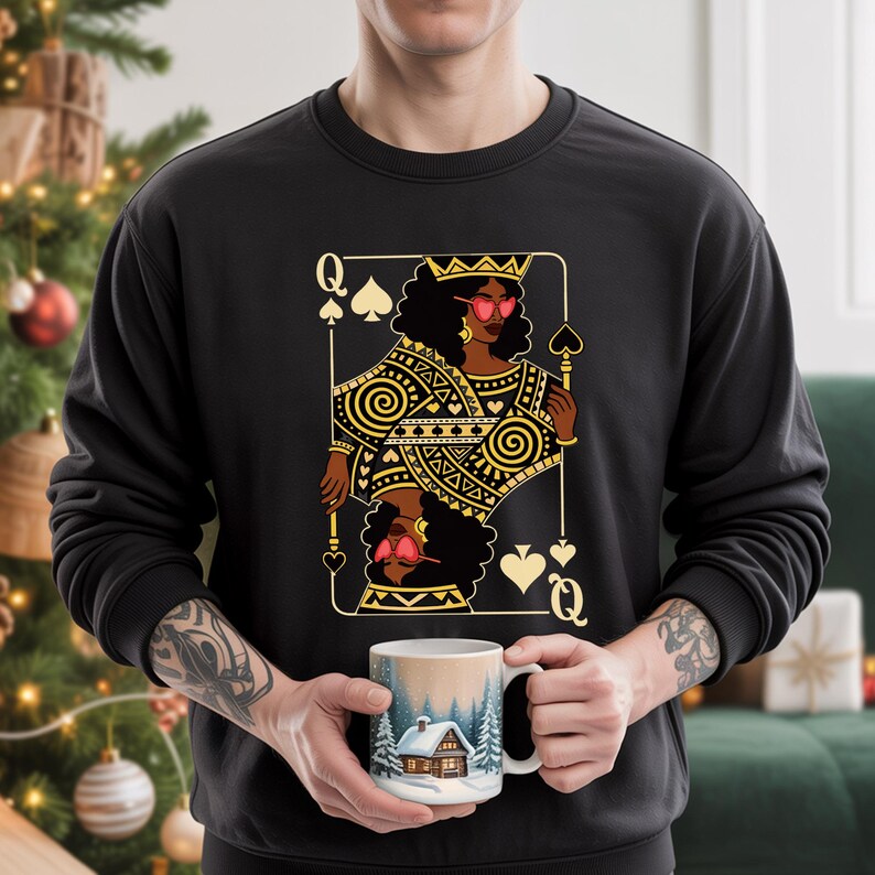 May include: Black sweatshirt with a gold and black playing card design featuring a queen wearing sunglasses. The person is holding a white mug with a winter scene.
