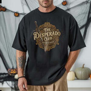 May include: Black t-shirt featuring a vintage-style graphic with the text "The Desperado Club" and the tagline "So Fun It Hurts." The design includes a sword and ornate scrollwork in shades of brown and gold. The shirt has a relaxed fit.