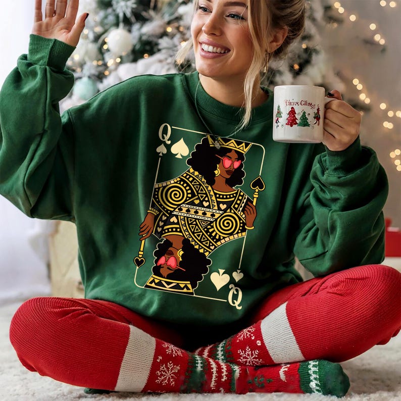 May include: A dark green sweatshirt with a Queen of Spades playing card design featuring a Black woman wearing sunglasses. The person is wearing red trousers and Christmas socks, and holding a white mug with a Christmas tree design.