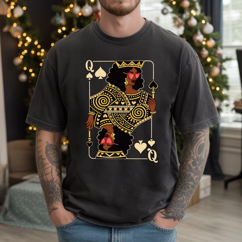 May include: Dark grey t-shirt with a gold and black Queen of Spades playing card design. The card shows a woman wearing sunglasses and a crown. The shirt is a casual, unisex style.