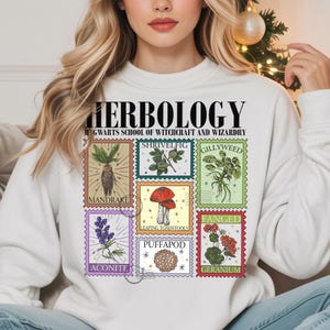 May include: White sweatshirt featuring a colorful graphic of various plants and the text "Herbology" and "Hogwarts School of Witchcraft and Wizardry". The graphic includes images of Mandrake, Shrivelfig, Gillyweed, Aconite, Leaping Toadstools, Puffapod, and Geranium.