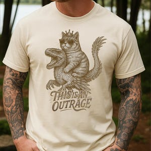 May include: Beige t-shirt featuring a detailed graphic of a cat wearing sunglasses and a crown, riding a dinosaur. The text "THIS IS AN OUTRAGE" is below the illustration. The shirt is made of a soft material.
