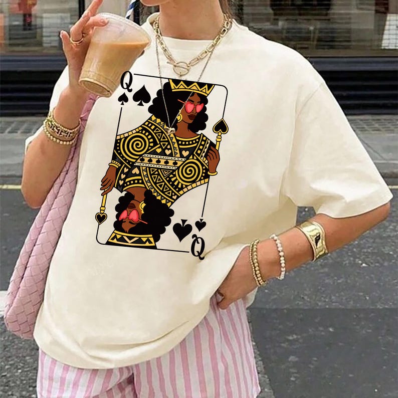 May include: Cream-coloured t-shirt featuring a playing card design of a queen of spades. The queen is depicted with black hair, a gold crown, and pink sunglasses. The shirt is paired with pink and white striped shorts.