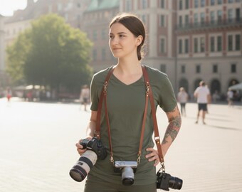 May include: A brown leather camera strap with three cameras attached. The strap is worn over the shoulders. The cameras are black and silver. The person is wearing a green top and trousers.