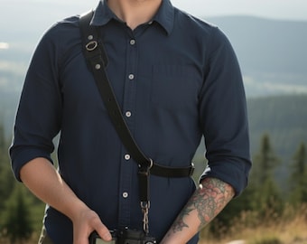 May include: Black leather camera strap with a shoulder pad and adjustable waist strap. The strap is attached to a DSLR camera. The photographer is wearing a dark blue button-down shirt.