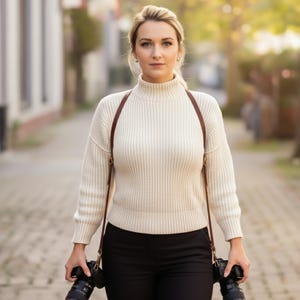May include: A woman wearing a cream-colored turtleneck sweater and black pants, holding two cameras. She has a brown leather camera strap across her shoulders. The background is a blurred street scene with buildings and trees.