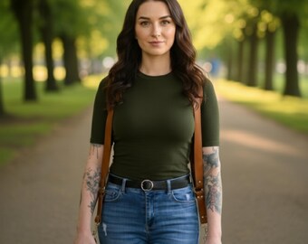 May include: A woman wearing a dark green short-sleeved shirt and blue jeans, with a brown leather camera strap. She is holding a camera and standing on a path lined with trees.