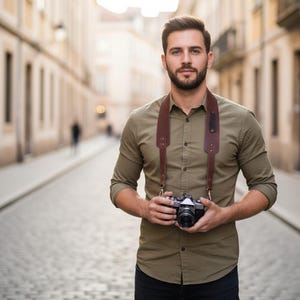 May include: A man wearing a green button-down shirt and black pants, holding a black and silver camera with a brown leather strap. The camera strap is around his neck. The background is a cobblestone street with buildings on either side.