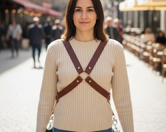 May include: A brown leather camera harness with metal rings and rivets. The harness is worn over a cream-colored sweater. Two cameras are attached to the harness. The background shows a city street with people and buildings.